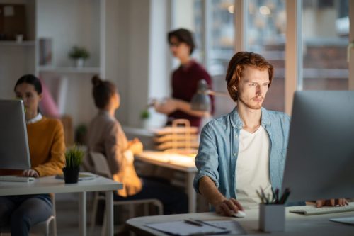 Young serious man in casualwear sitting in front of computer by desk and looking through websites in working environment